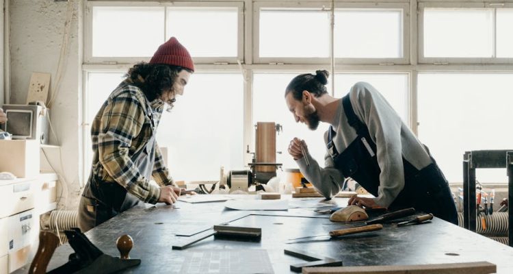 Two male carpenters in a workshop discuss a project, surrounded by tools and wood.